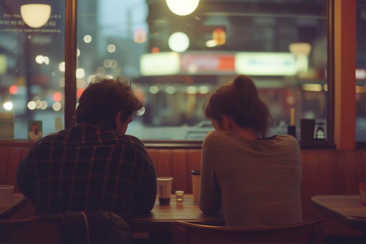 Couple in a dimly lit diner, backs turned and heads bowed, crafting a plan in hushed tones, overlooking urban street life at dusk through towering glass walls